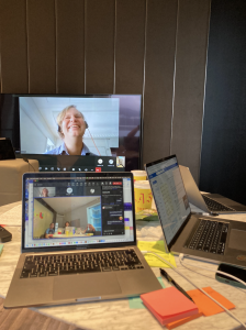jesper laughs on a large TV screen - the rest of the meeting is visible through the three laptops clustered around the larger TV. Post-it notes and pens are on the table.